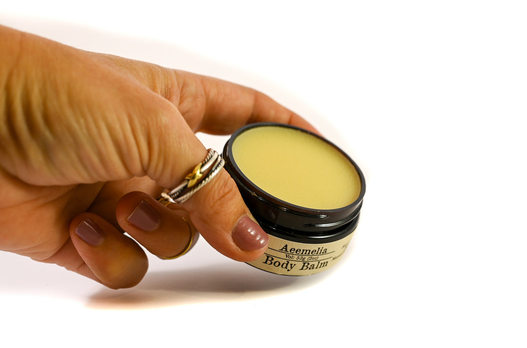 Aeemelia Body Balm, a rich, concentrated moisturizer that utilizes the nutrients of shea butter and beeswax to heal dry skin. Black screw top lid on 2 oz amber plastic jar with white and black label.  Suitable for all skin types (including sensitive skin). With hand to so relative size of container. 