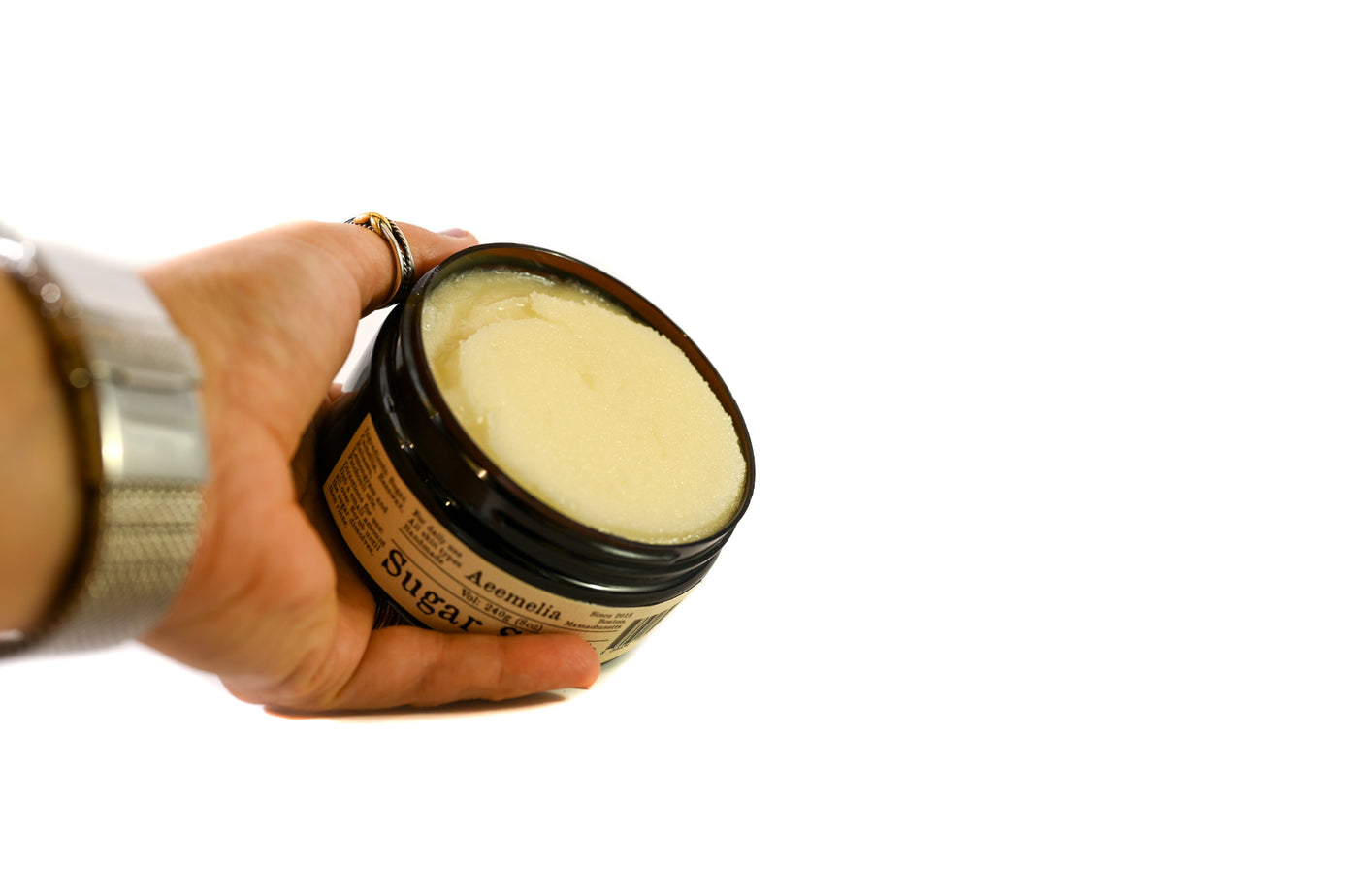 Aeemelia sugar scrub’s unique texture is packed with the nutrients of camellia seed oil and babassu oil to perfectly hydrate your skin. Black screw top lid on 16 oz or 8 oz amber plastic jar with white and black label.  Suitable for all skin types (including sensitive skin). With hand to show size relative size of the jar. 
